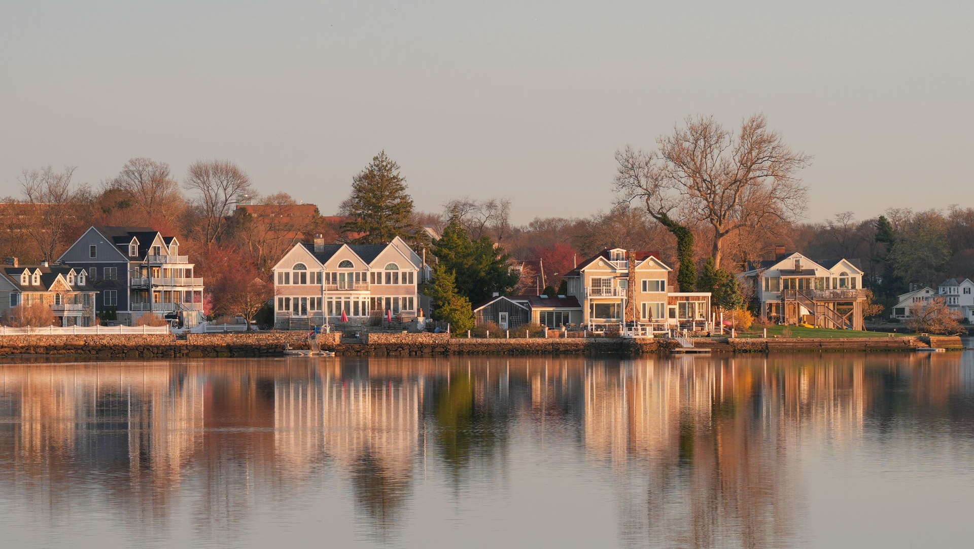 Row of waterfront houses at sunset reflected in calm water with autumn trees behind them