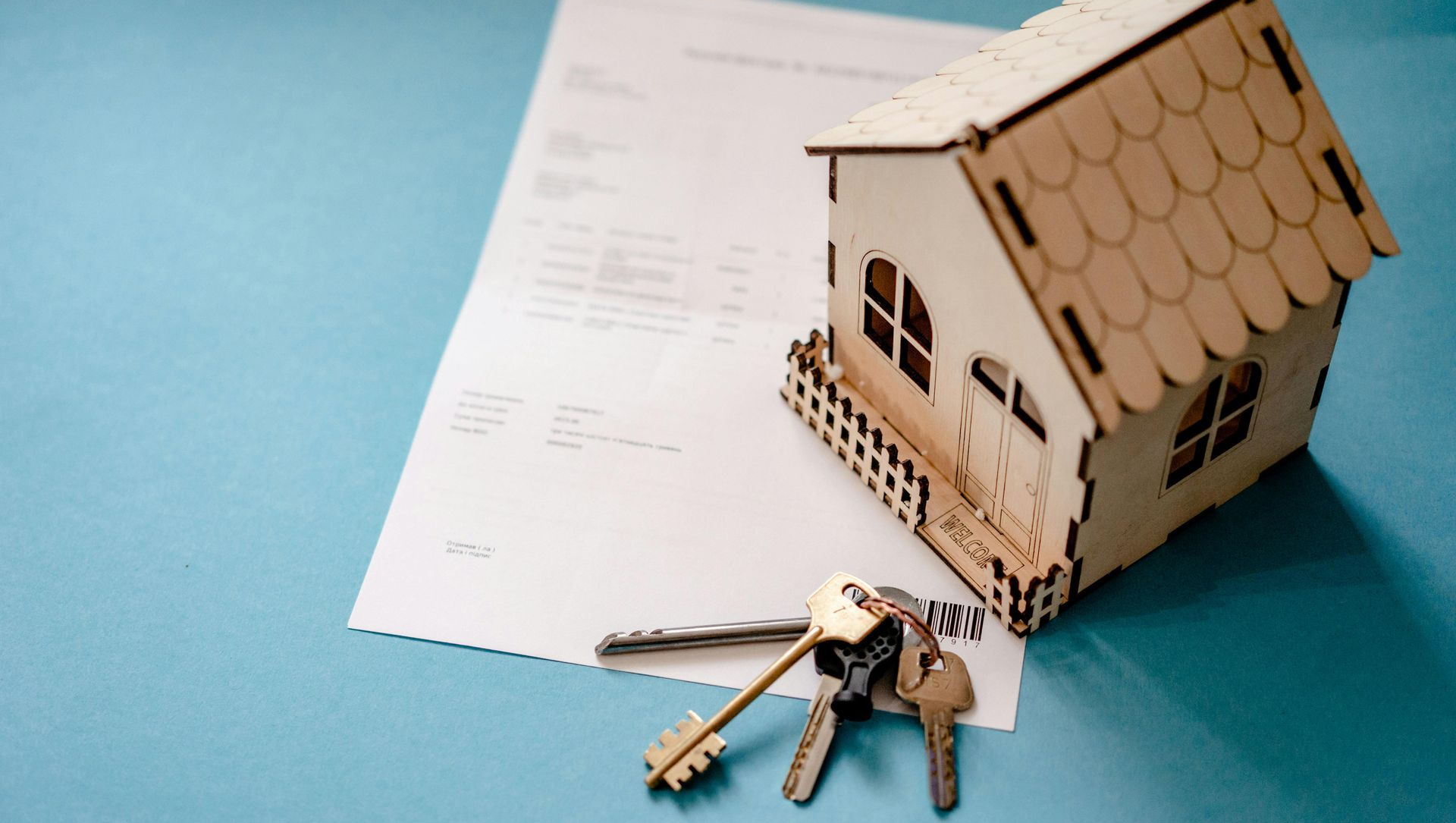 A small wooden house model and a cluster of metal keys sit on a document against a solid blue background.