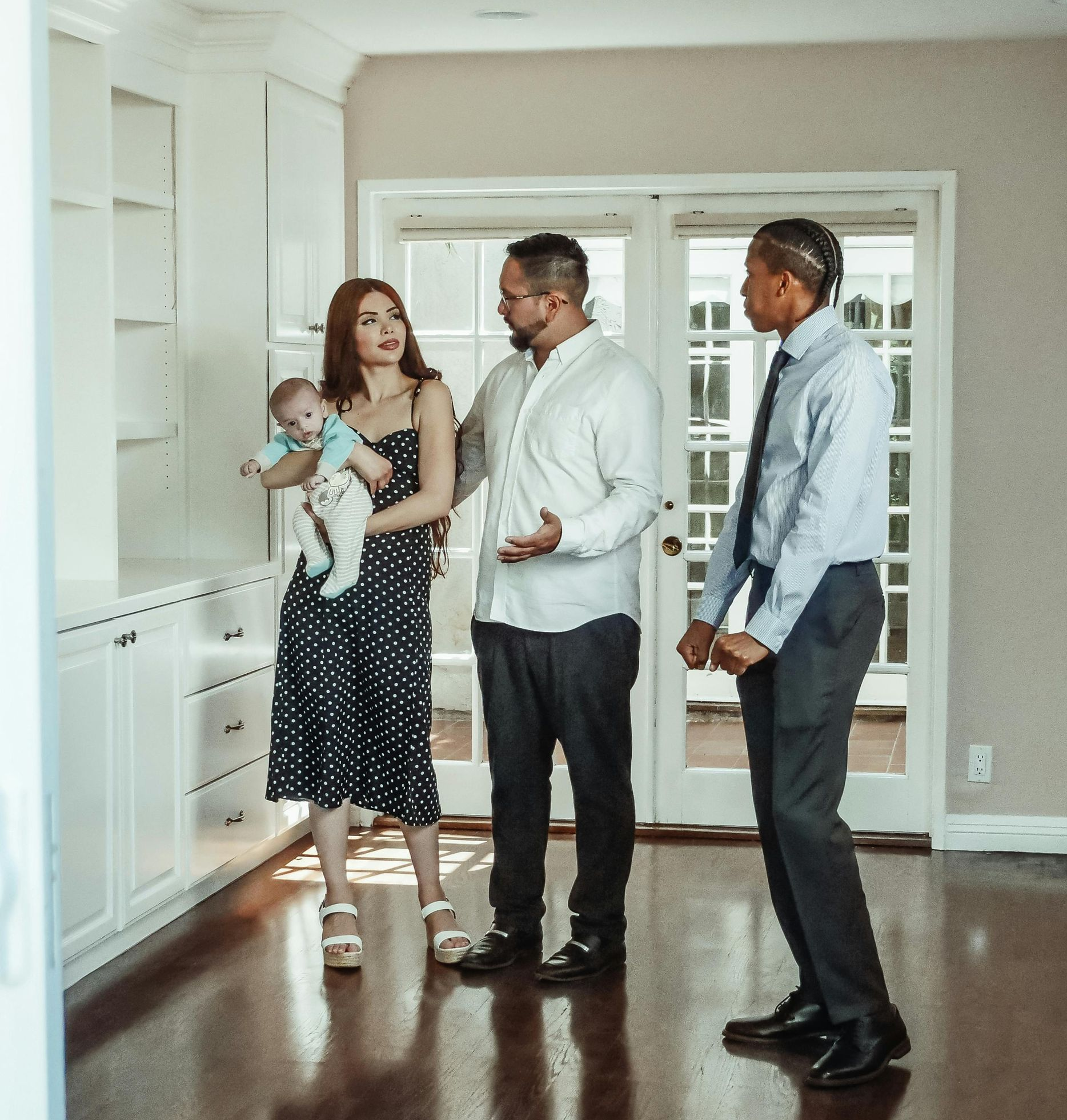 Family with realtor viewing a house. Baby held by woman, two men talking near open doors.