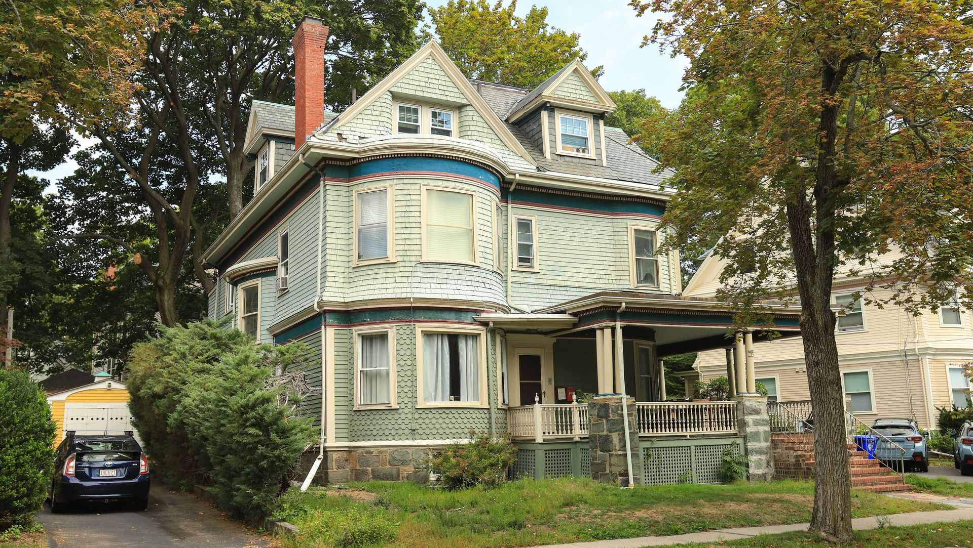 Old pale-blue Victorian house with porch, trees, and parked cars on a residential street