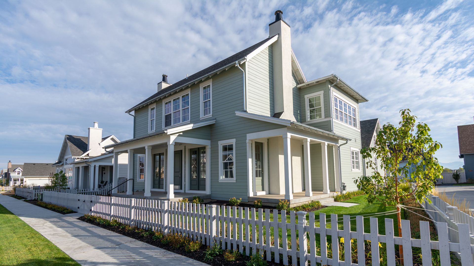 Gray two-story house with white trim and picket fence along a sunny sidewalk.