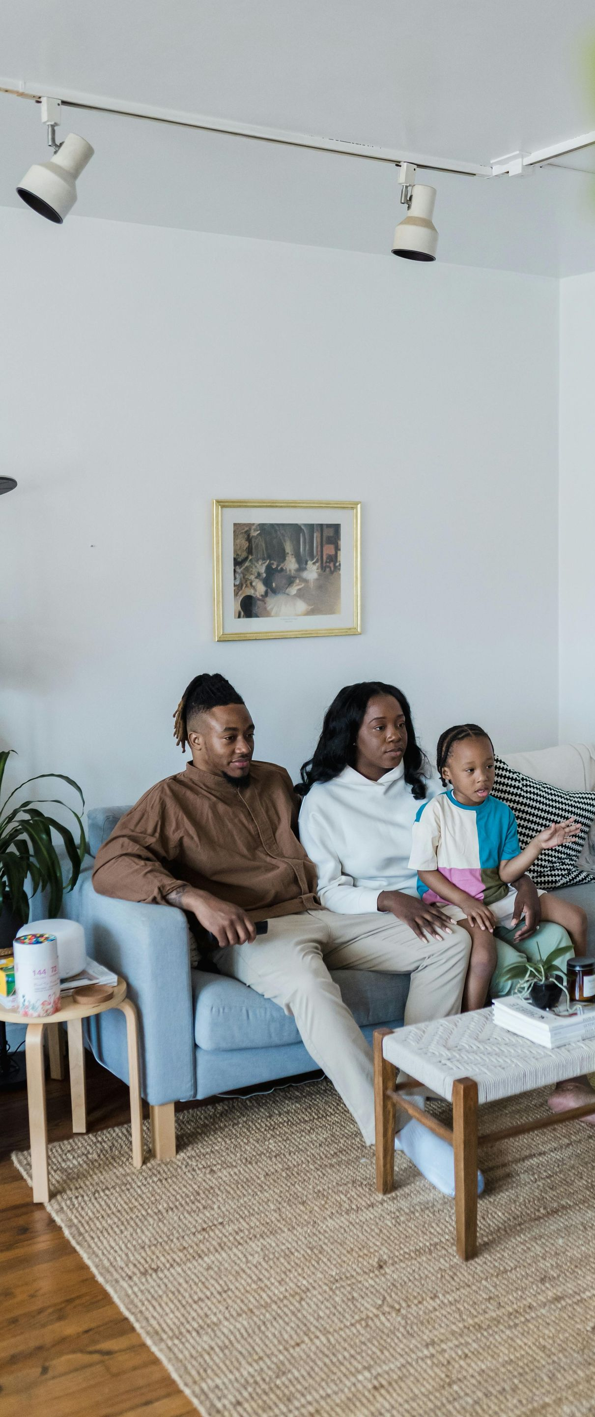 Three people sit on a light blue sofa in a bright living room, with a coffee table and plants nearby.