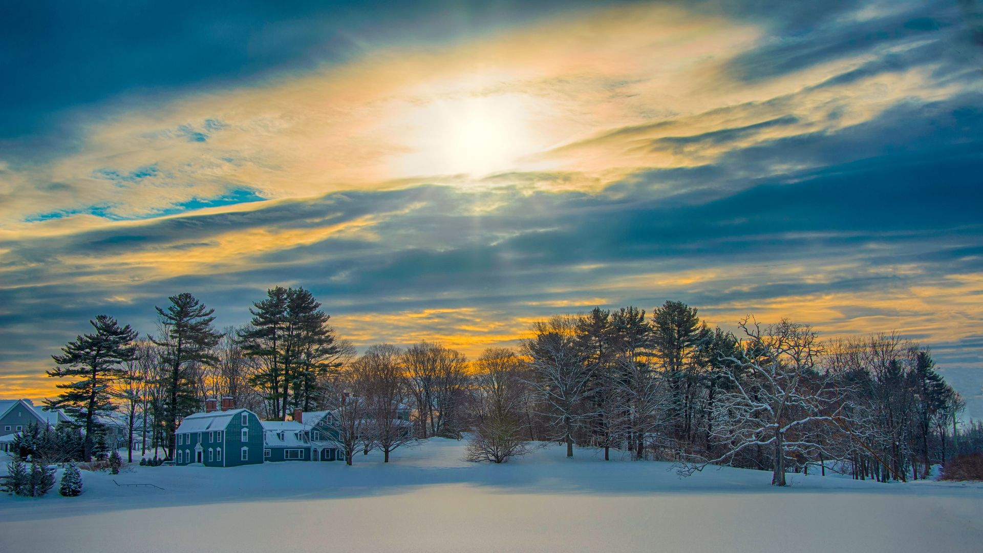 Snow-covered field at sunset with a house, bare trees, and a glowing cloudy sky