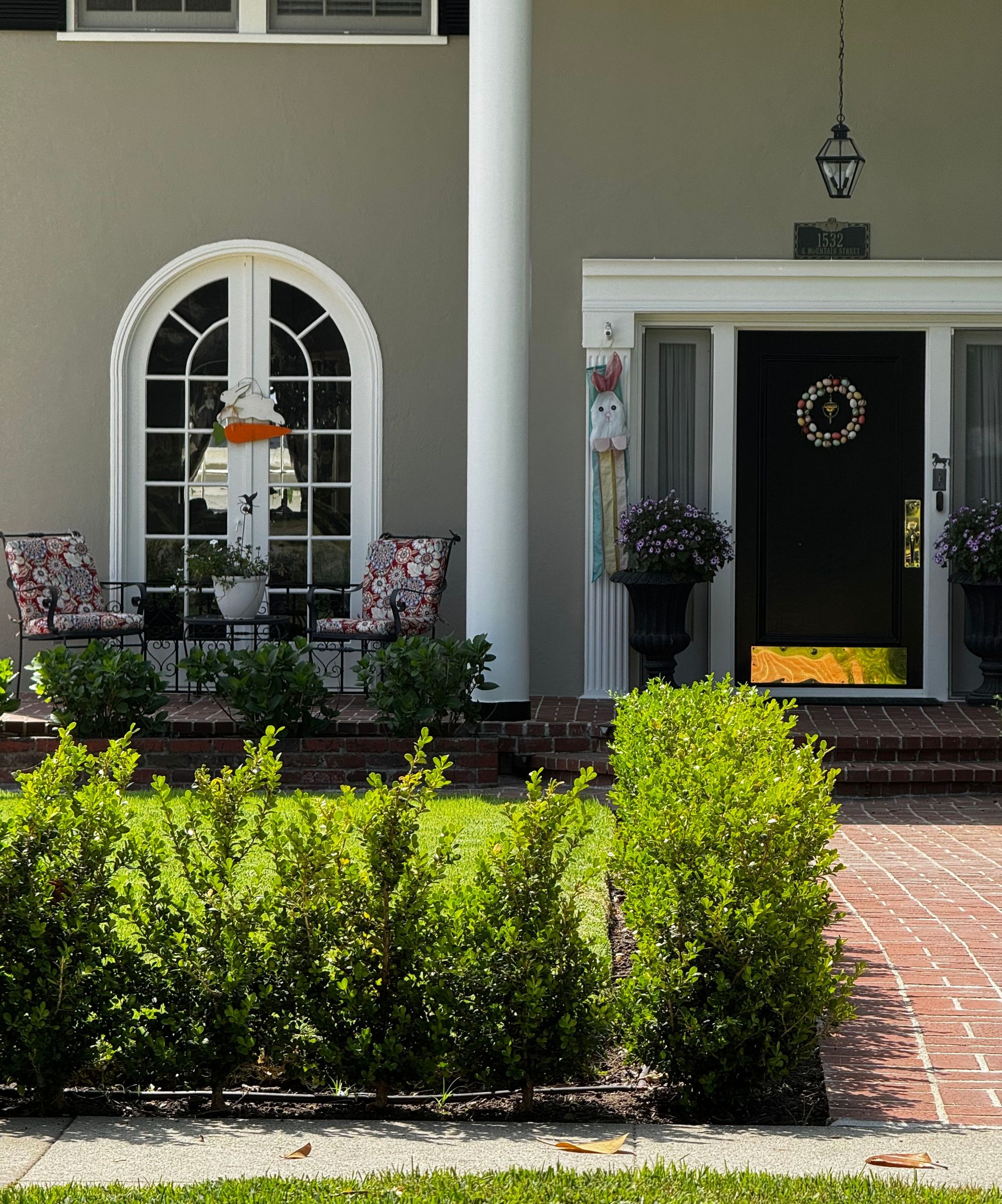 Front porch with arched window, white door, potted flowers, and trimmed green shrubs by a brick walkway
