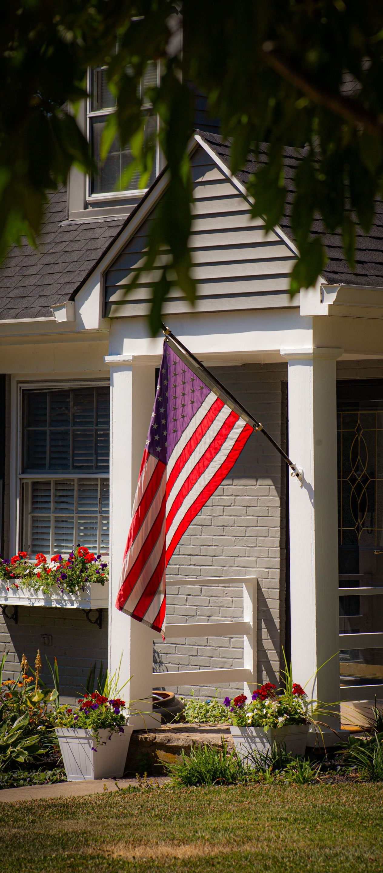 American flag hanging on the porch of a white house with potted flowers and shaded greenery