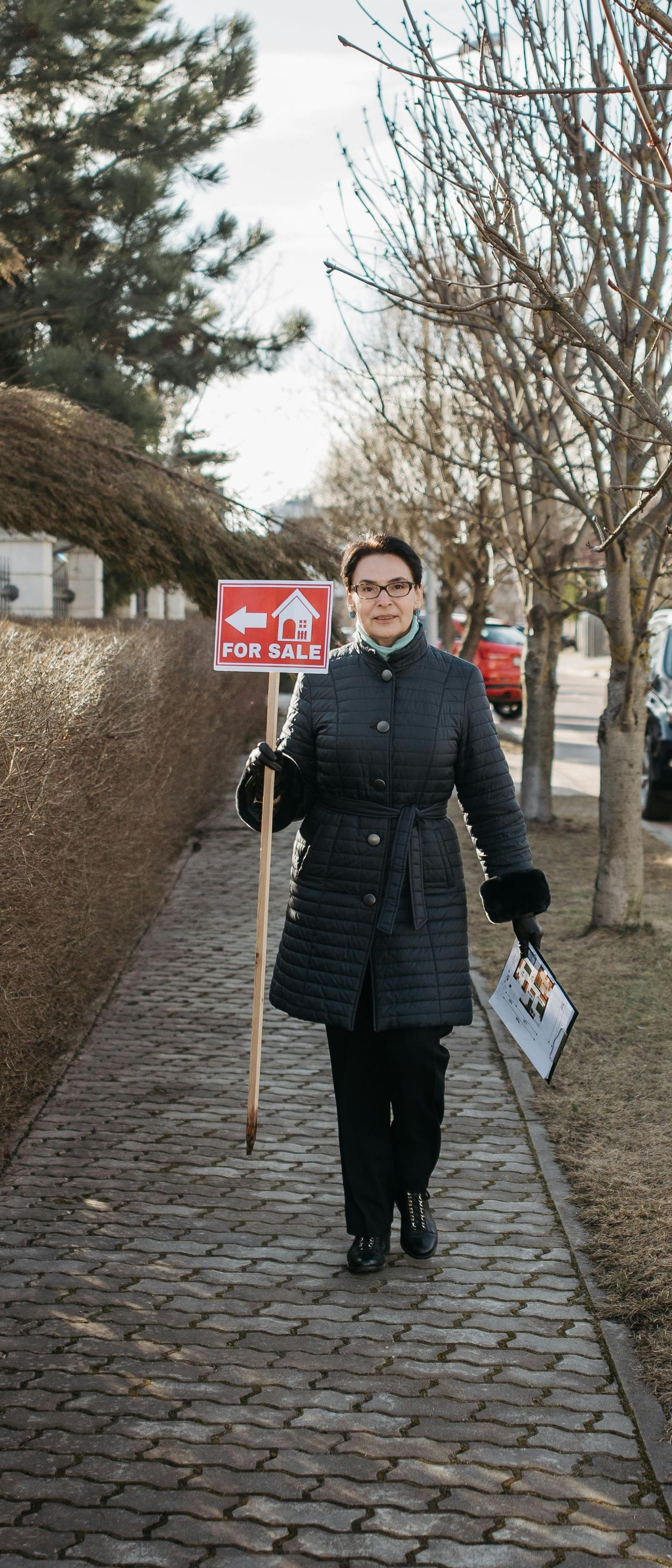 Person holding a sign on a cobblestone sidewalk beside hedges and parked cars