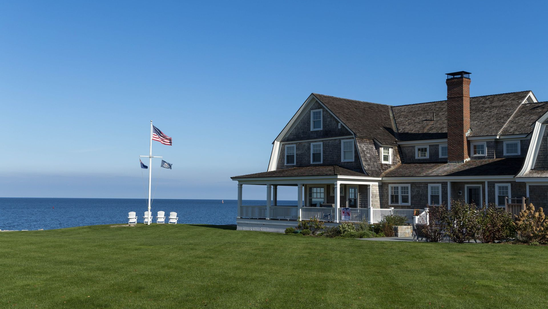 Coastal house with a flagpole on a grassy lawn beside the ocean under a clear blue sky