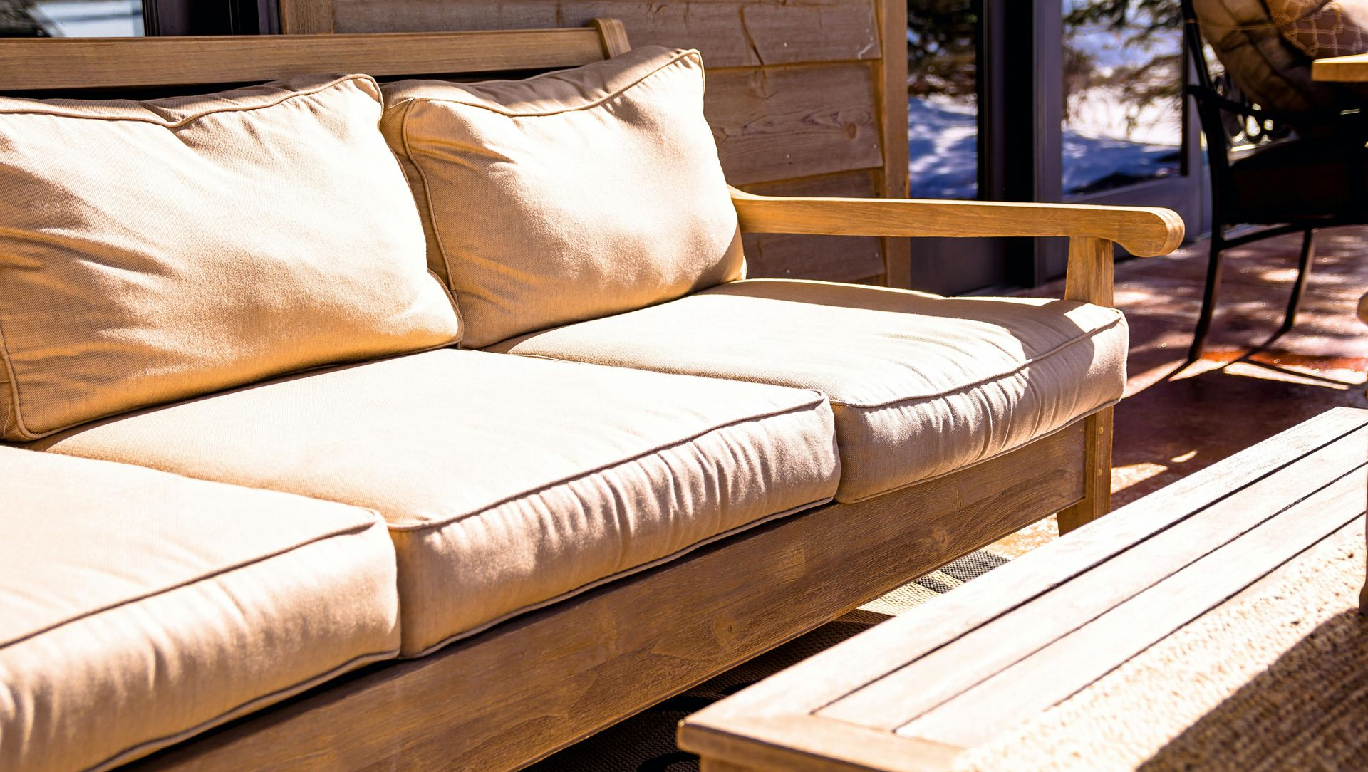 Wooden outdoor sofa with beige cushions on a sunlit deck beside a low table