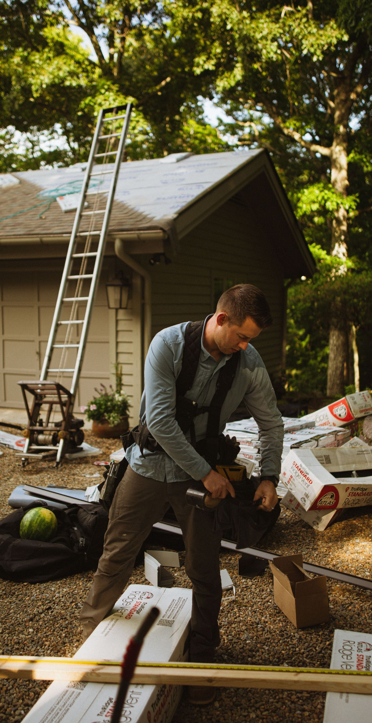 Man working outdoors beside a ladder and shed, assembling materials in a sunny yard.