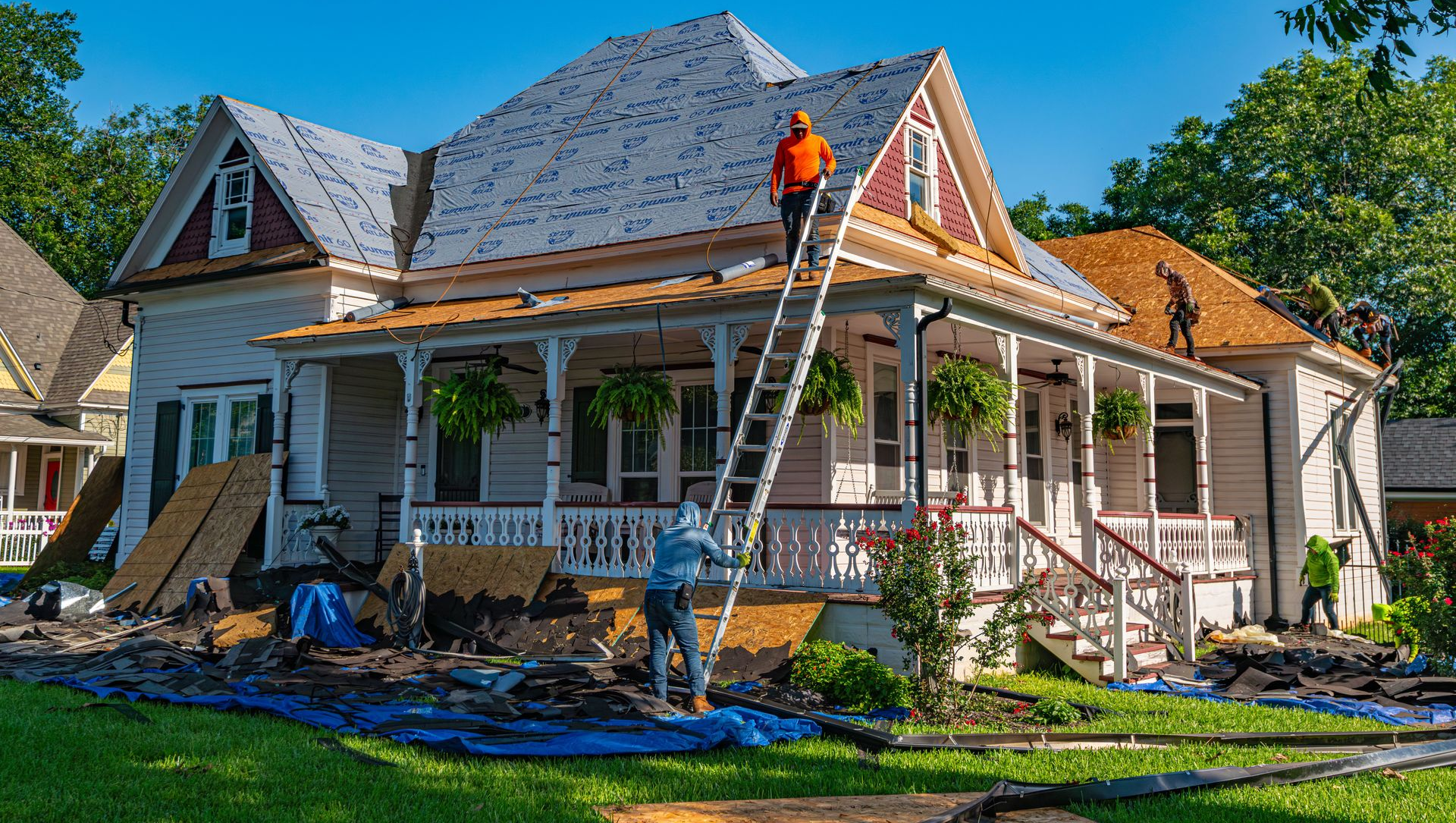 Workers repairing a house roof, with ladders, tools, and tarps around the porch and yard.