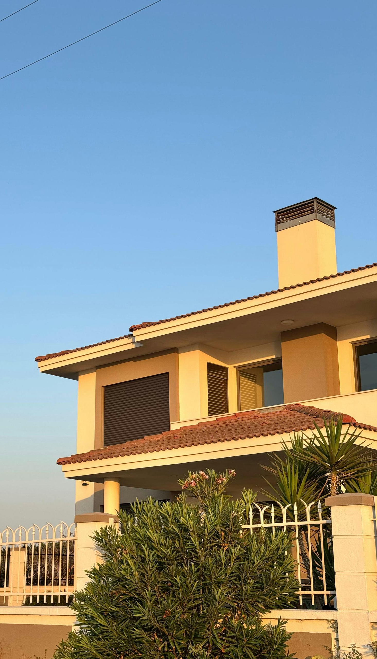 Sunlit two-story house with cream walls, red roof, and a front garden under a clear blue sky