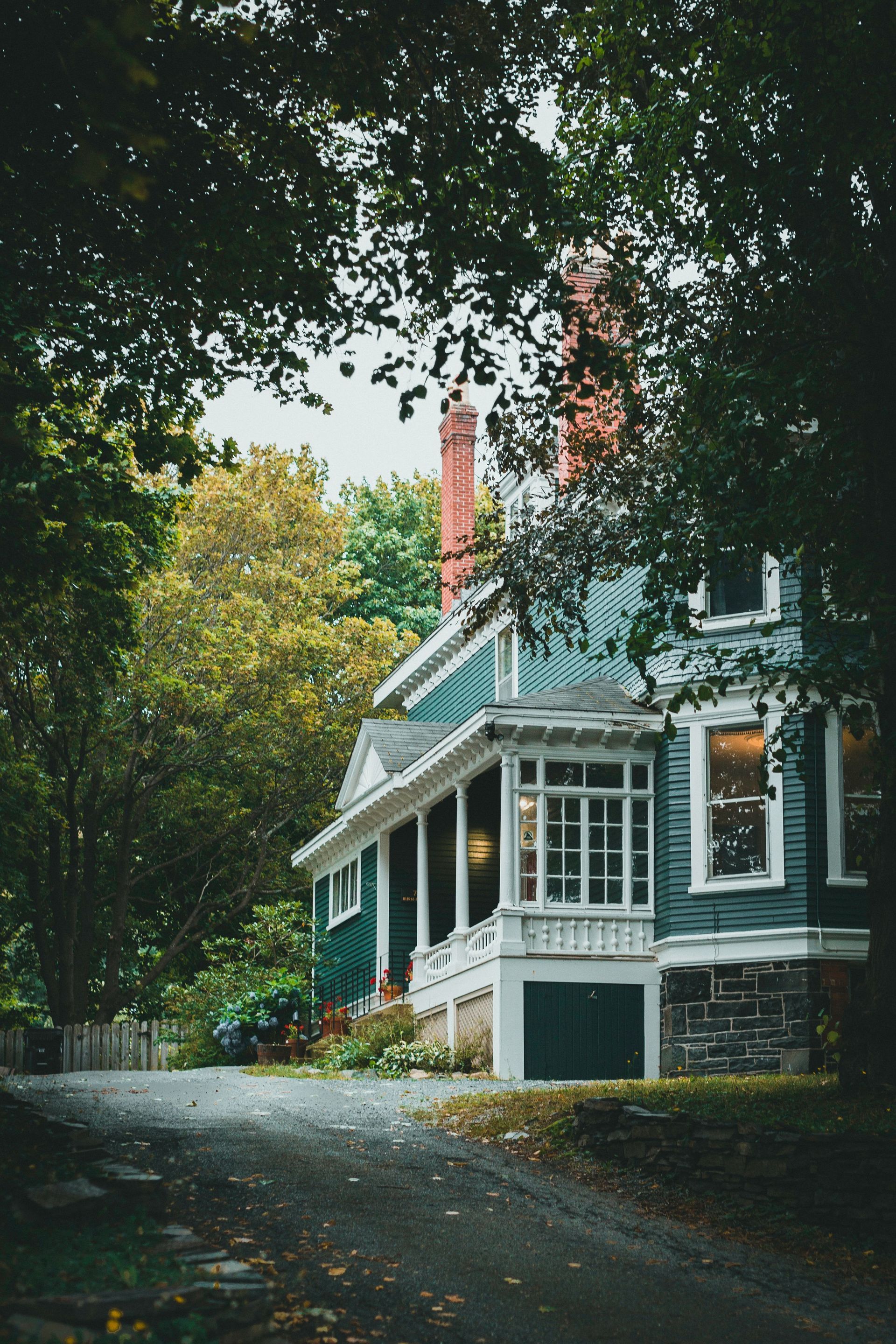 Victorian house with teal trim beside a wooded road, framed by overhanging trees