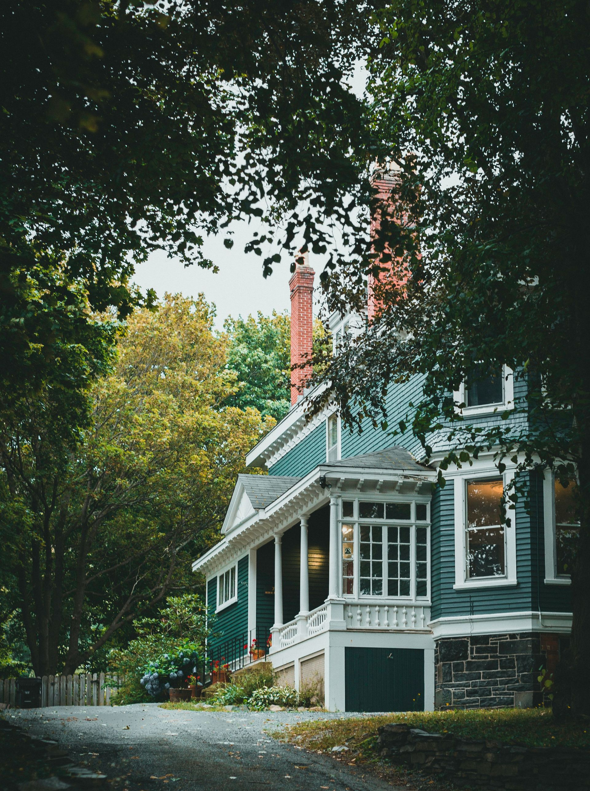 Tree-shaded blue house with white trim and a red chimney on a quiet street
