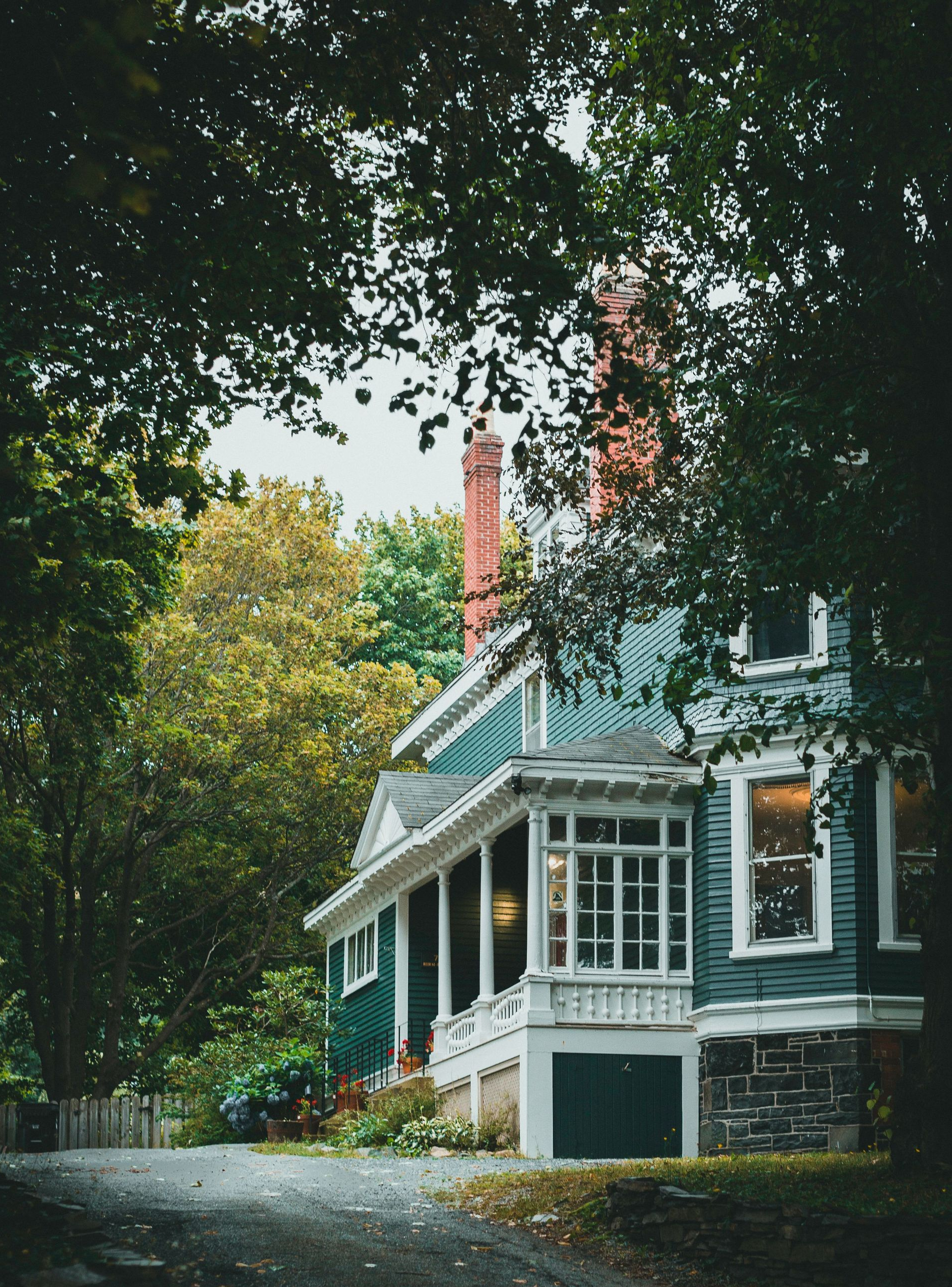 Tree-shaded blue house with white trim and a red chimney on a quiet street