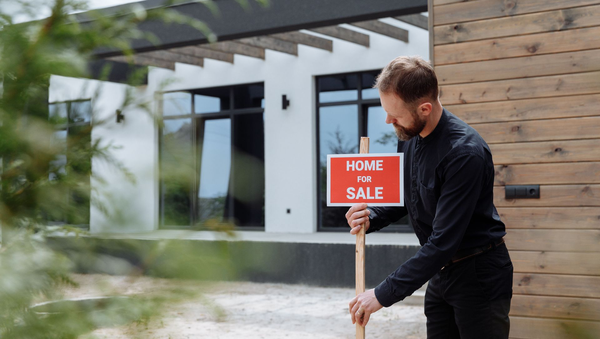 Man placing a red “Home for Sale” sign in front of a modern house