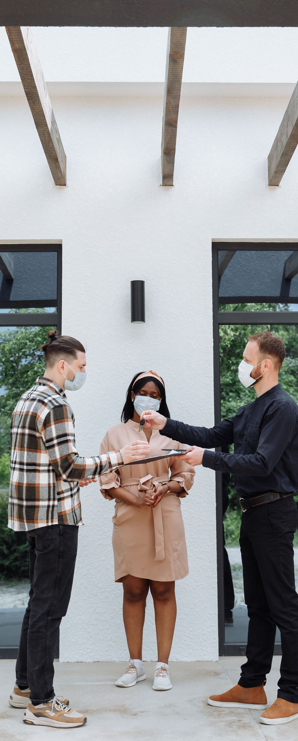 Three people wearing masks stand outdoors in front of a white building, holding and gesturing at a phone.