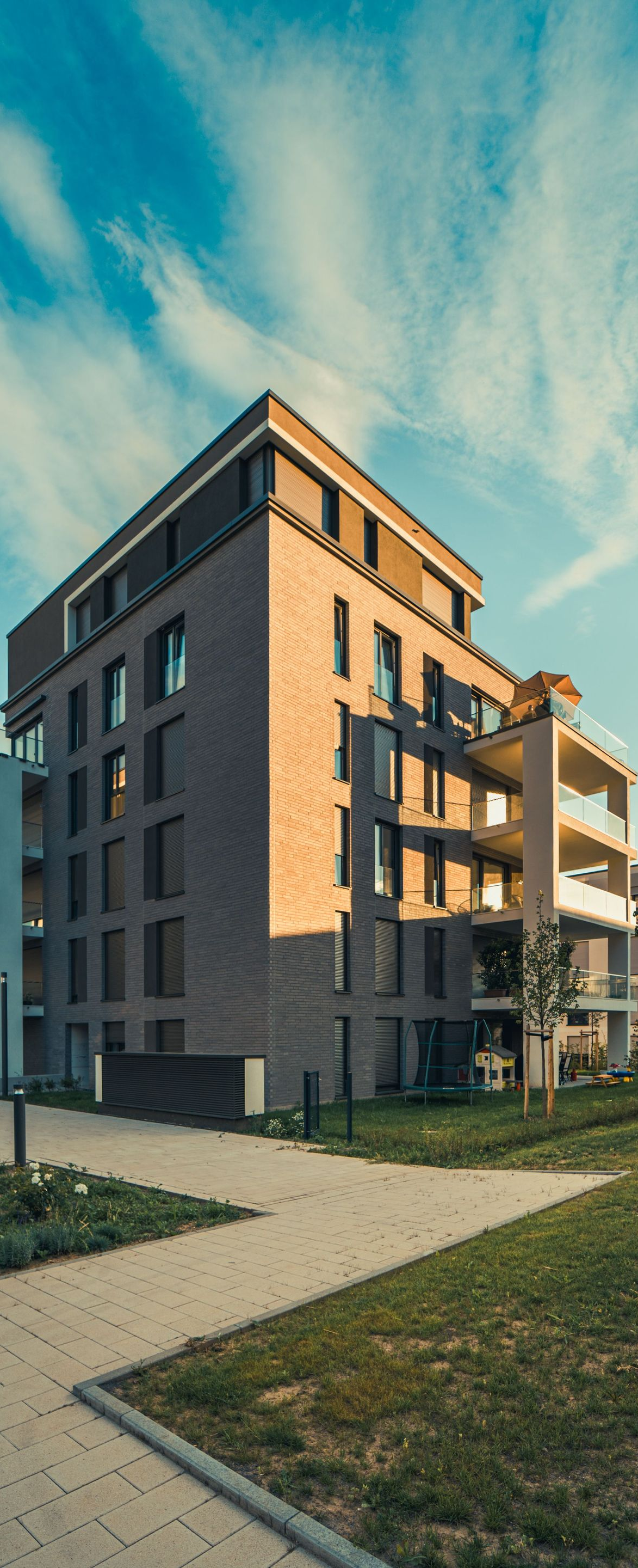 Modern apartment building with brick and white balconies beside a grassy path under a blue sky