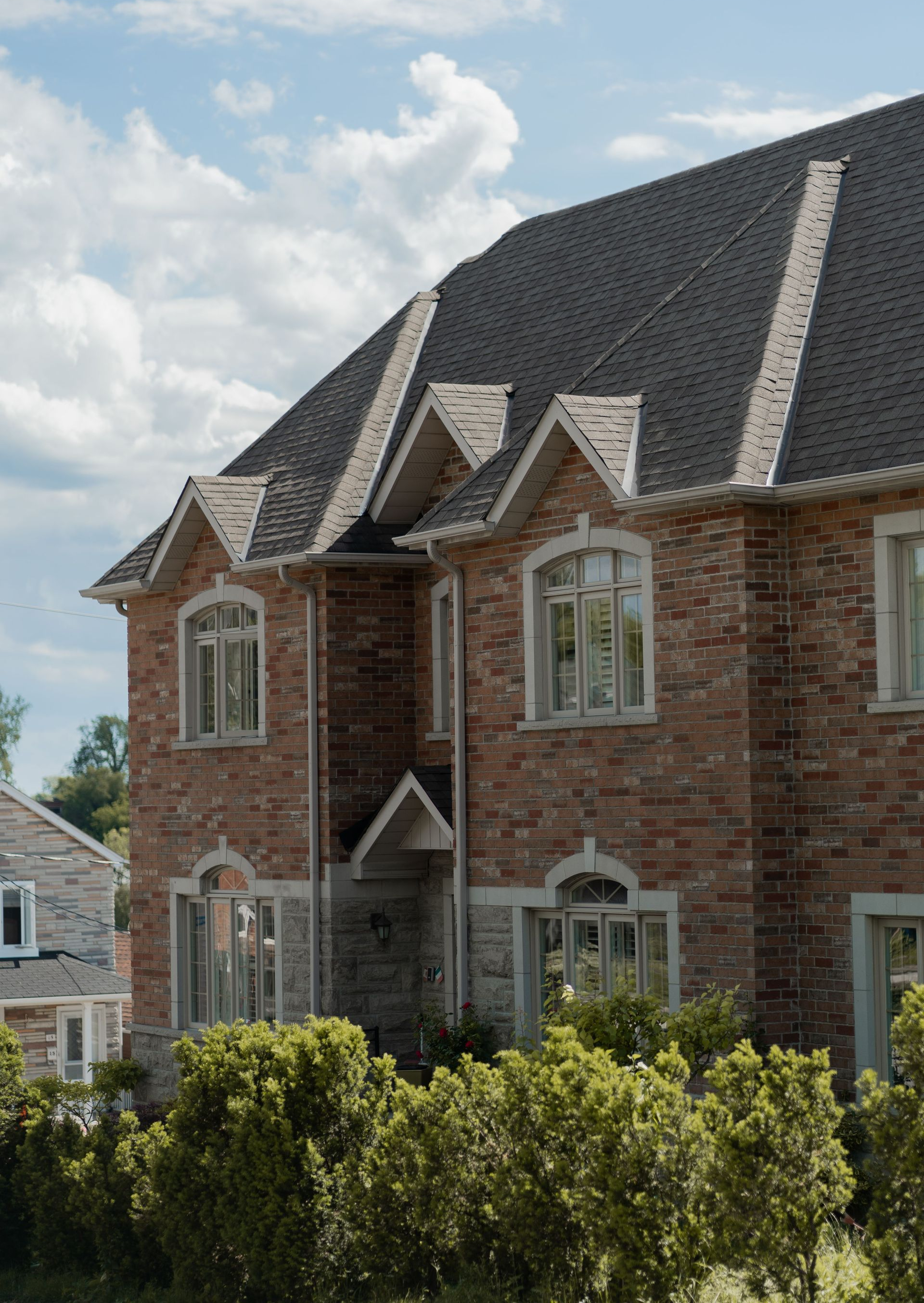 Brick townhouse with steep dark roof and arched windows behind green shrubs under a partly cloudy sky