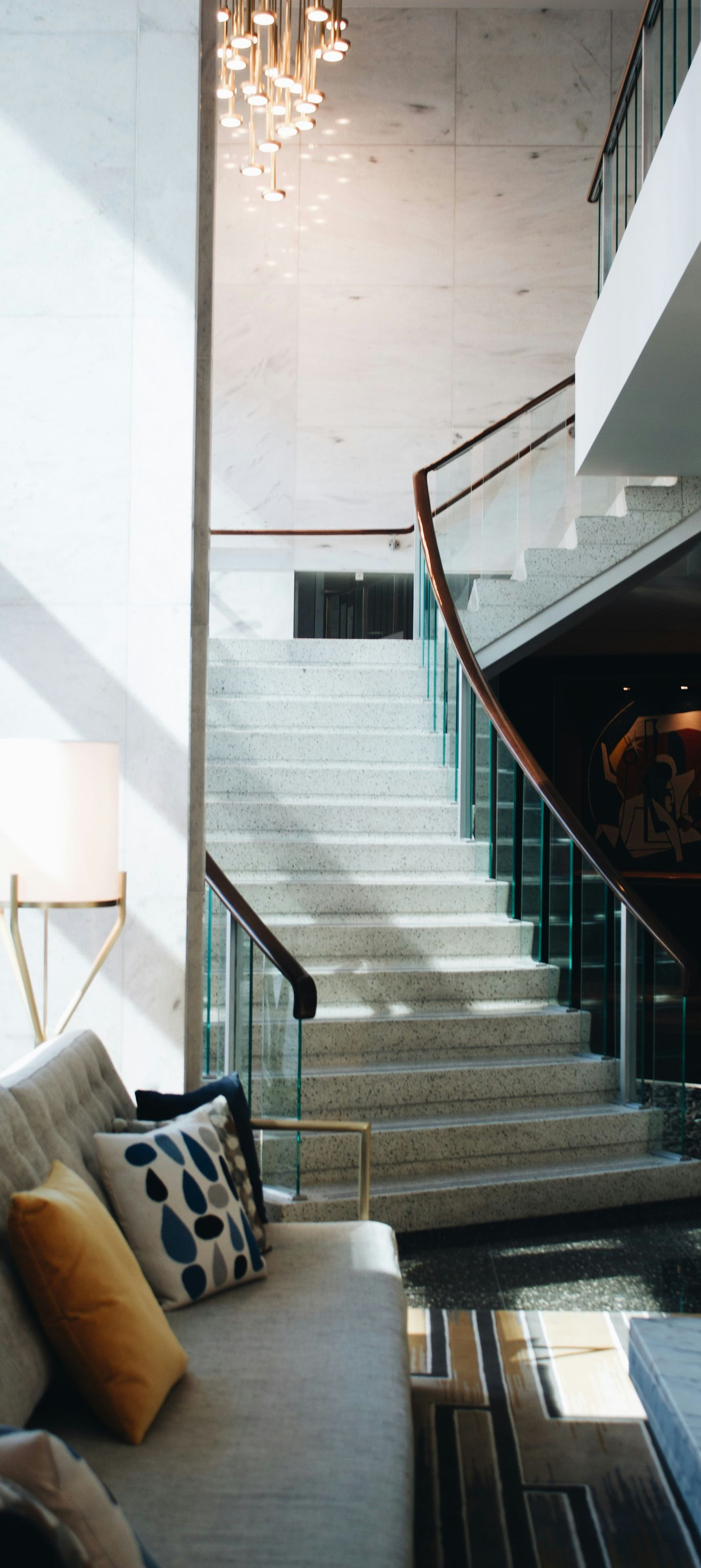 A staircase with stone steps and glass railings ascends toward a light-filled ceiling, viewed from a modern living room.