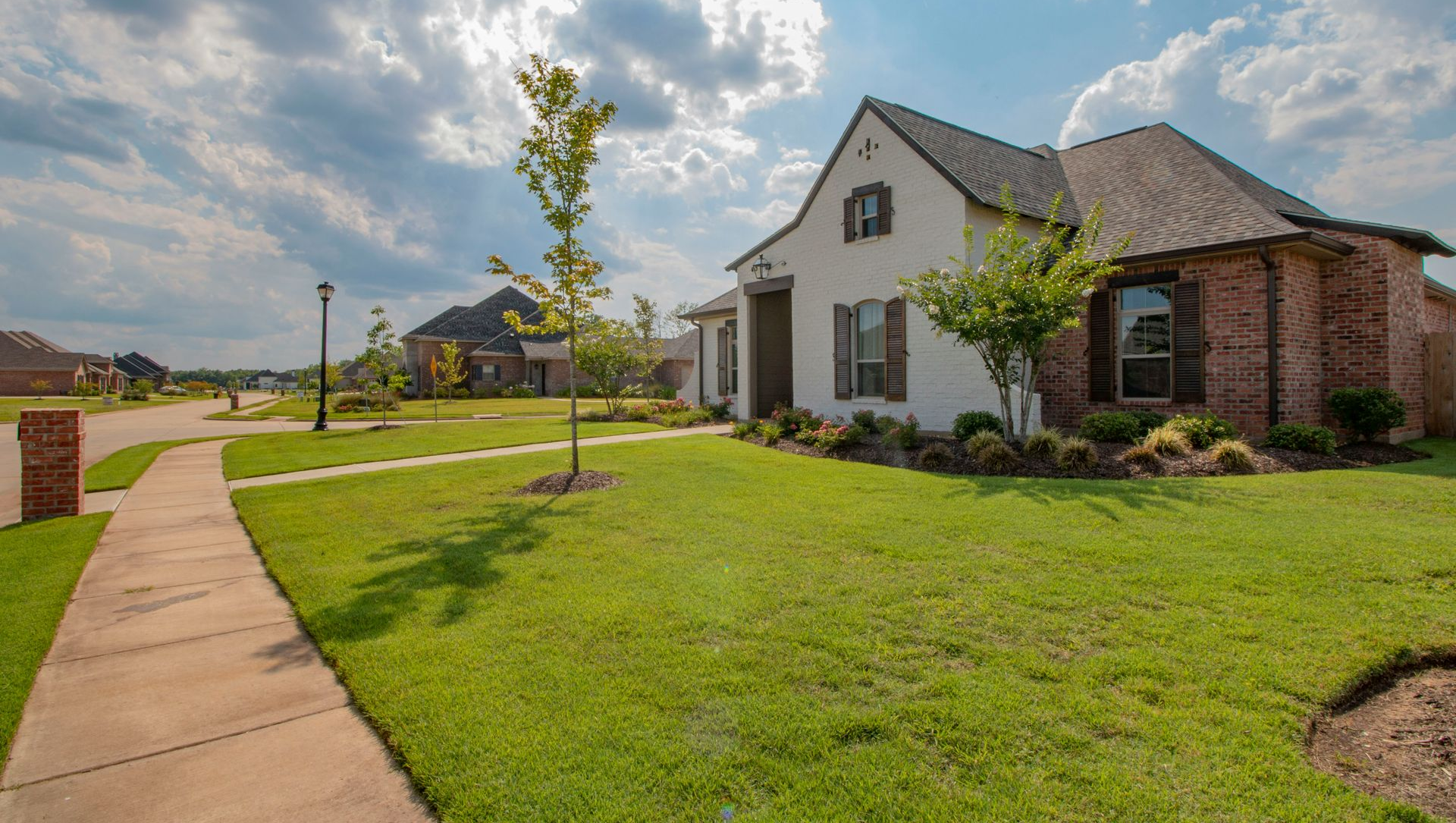 A beige brick house with a dark roof sits on a well-manicured lawn under a cloudy blue sky.