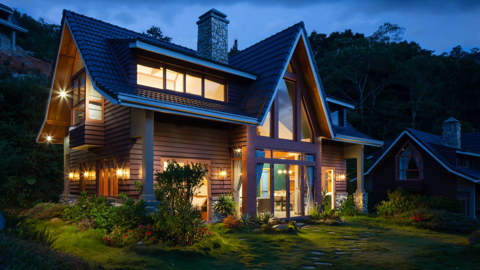 A two-story wood-paneled house with a steep, dark roof is illuminated by warm interior lights at dusk.