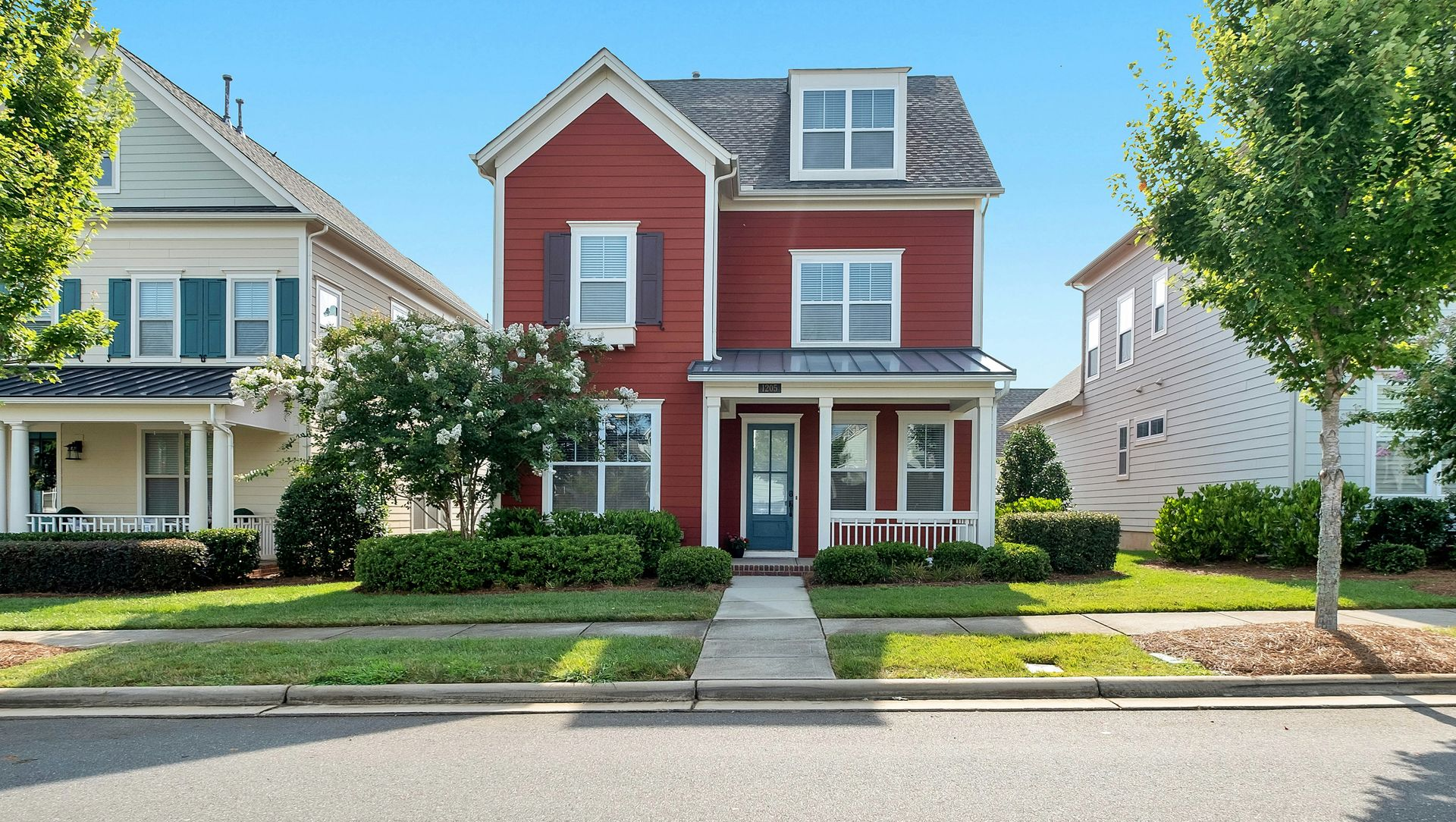 A red suburban house stands between two lighter-colored houses on a sunny street with green trees and lawns.