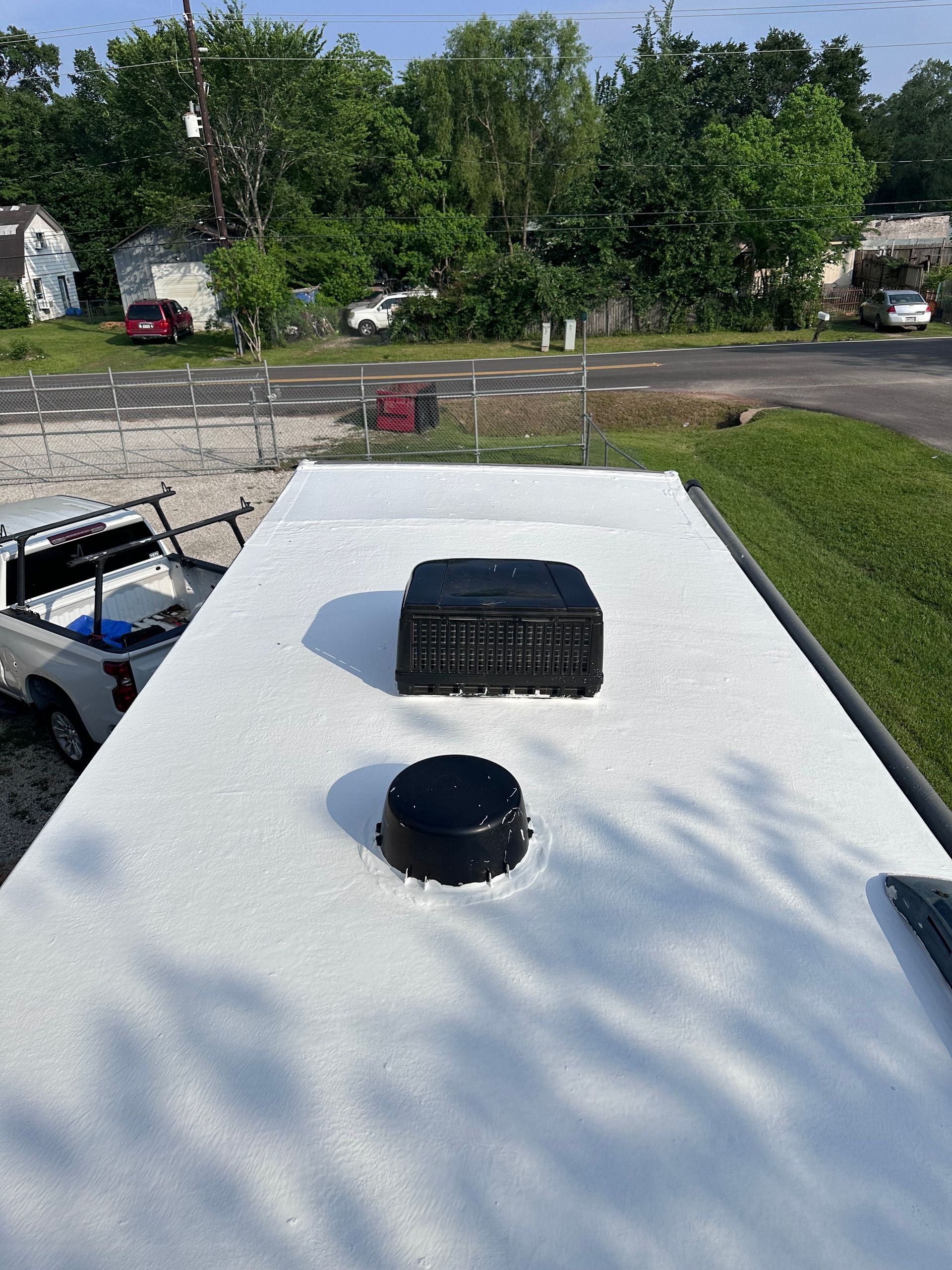 White RV roof with black vents in a sunny wooded parking area, with a golf cart at left.