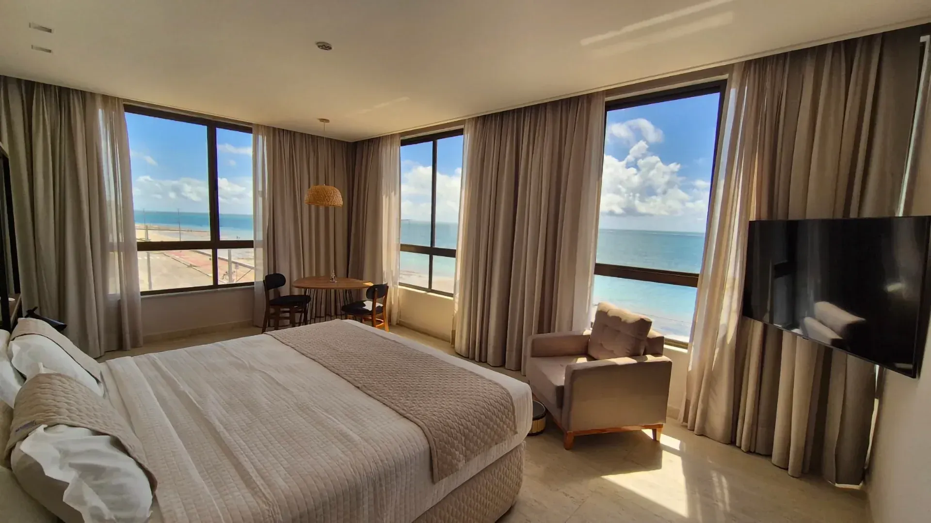 Quarto de hotel luminoso com cama, cadeira, TV e grandes janelas com vista para o oceano e o céu.
