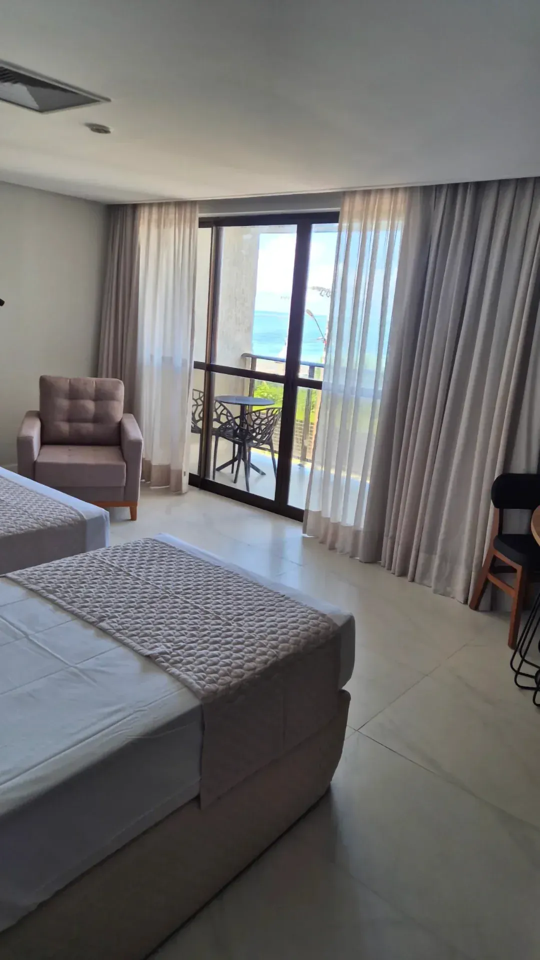 Quarto moderno com cama, poltrona, portas de varanda, cortinas e muita luz natural.