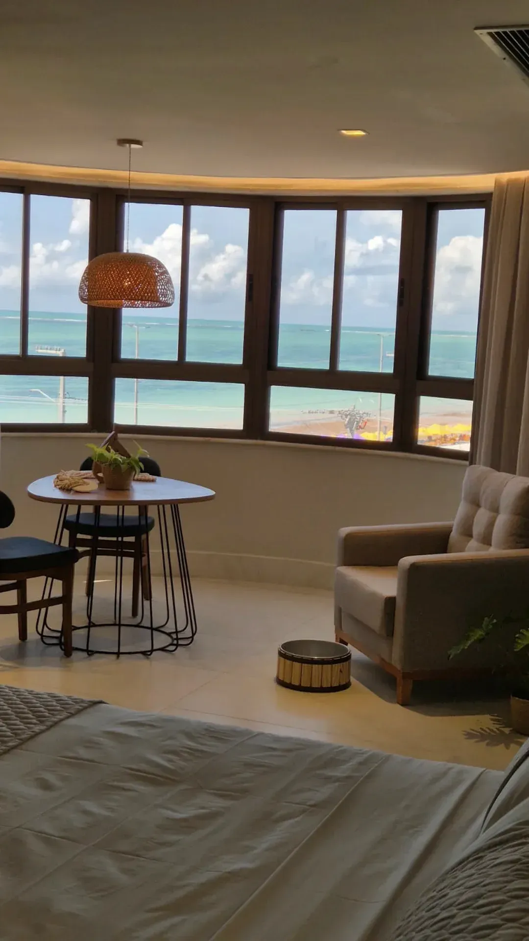 Quarto de hotel luminoso com janelas com vista para o mar, cama em primeiro plano, poltrona, mesa redonda.