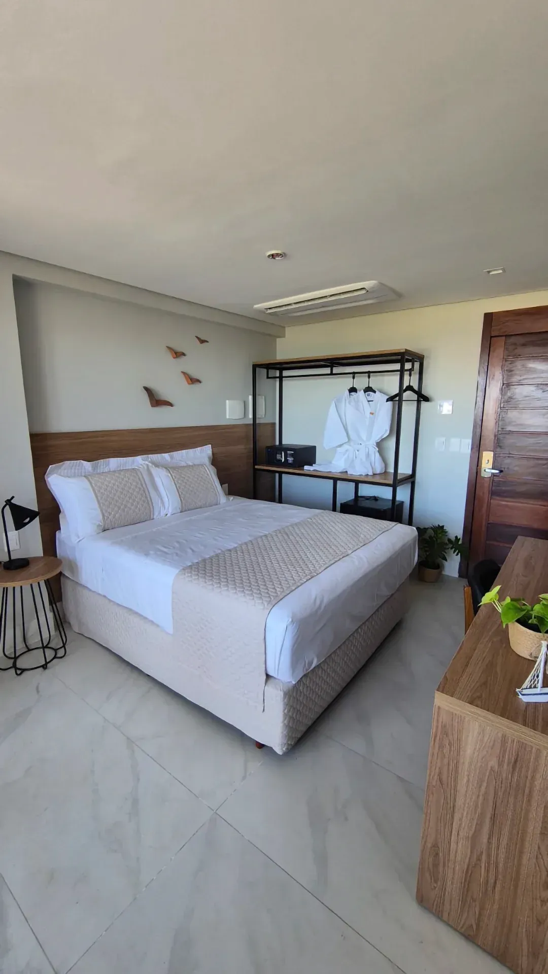 Quarto moderno e luminoso com cama, mesa de cabeceira, cômoda e guarda-roupa aberto com roupas penduradas.