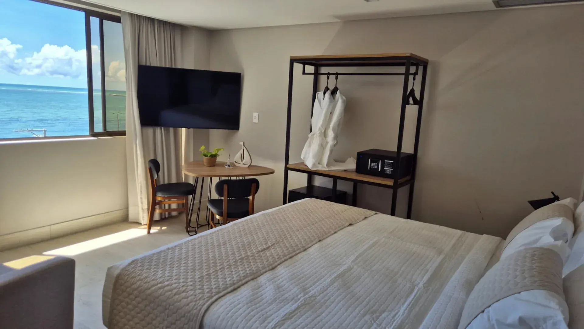 Quarto de hotel luminoso com cama, janela com vista para o mar, TV, mesa e cabide para roupas.