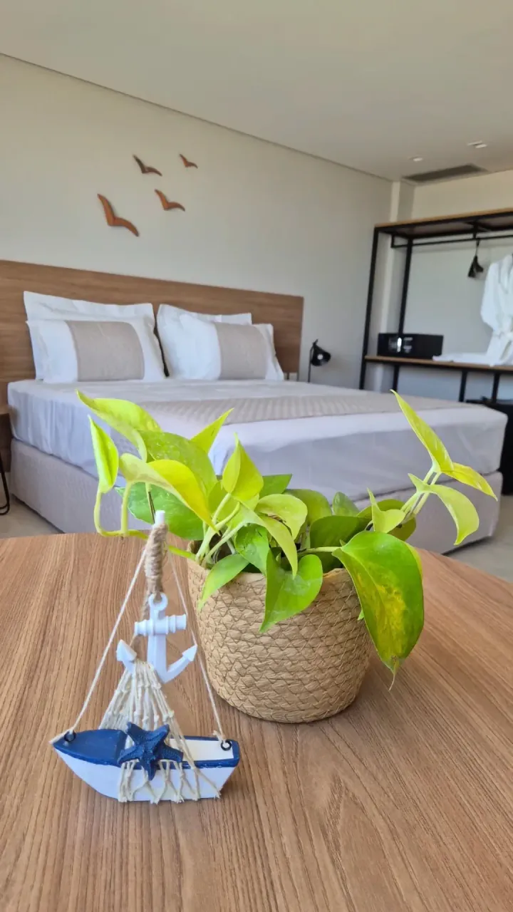 Quarto com tema de praia, com cama branca, mesa de madeira e planta em vaso em primeiro plano.