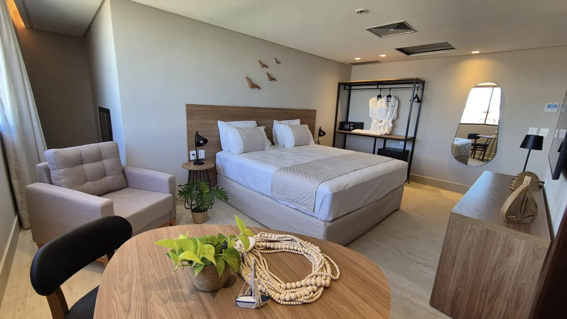 Quarto moderno e aconchegante com cama arrumada, poltrona, mesa redonda e espelho de parede em tons neutros suaves.