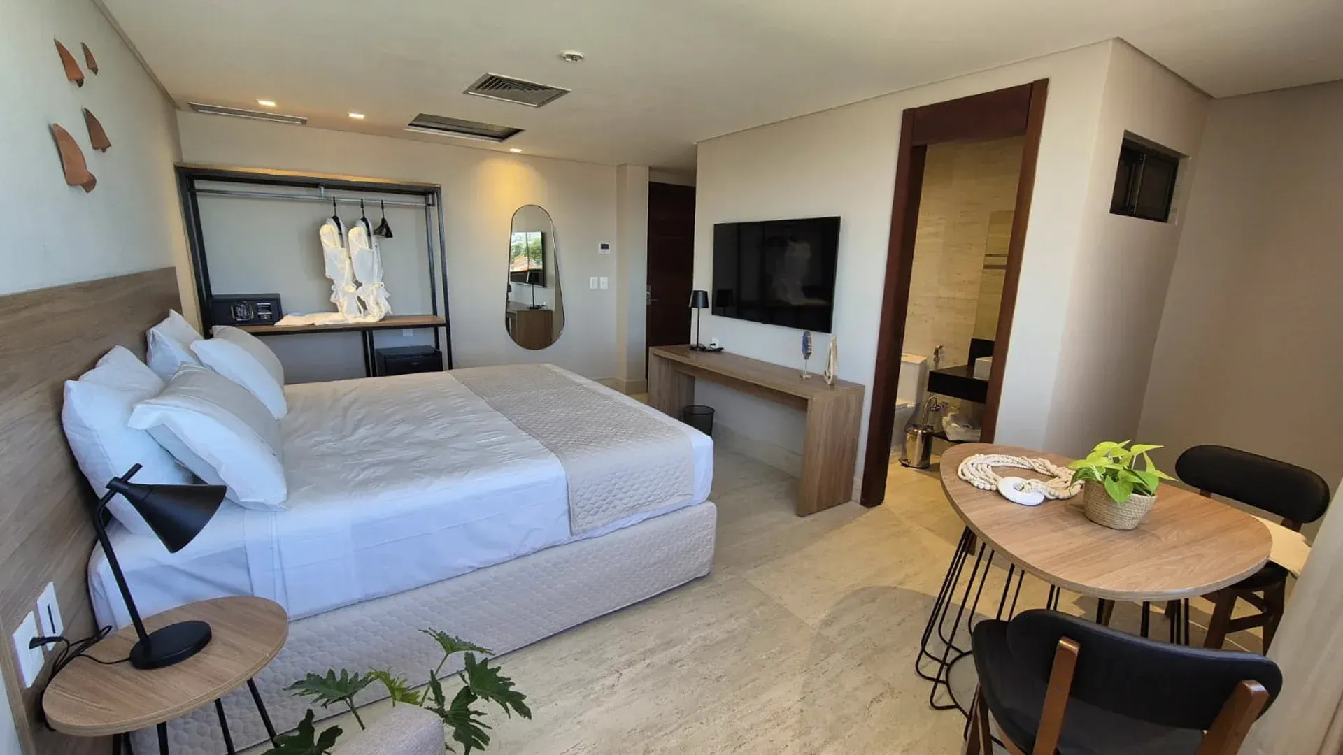 Quarto estúdio moderno com cama, TV, mesa de jantar e cozinha compacta em tons neutros e aconchegantes.