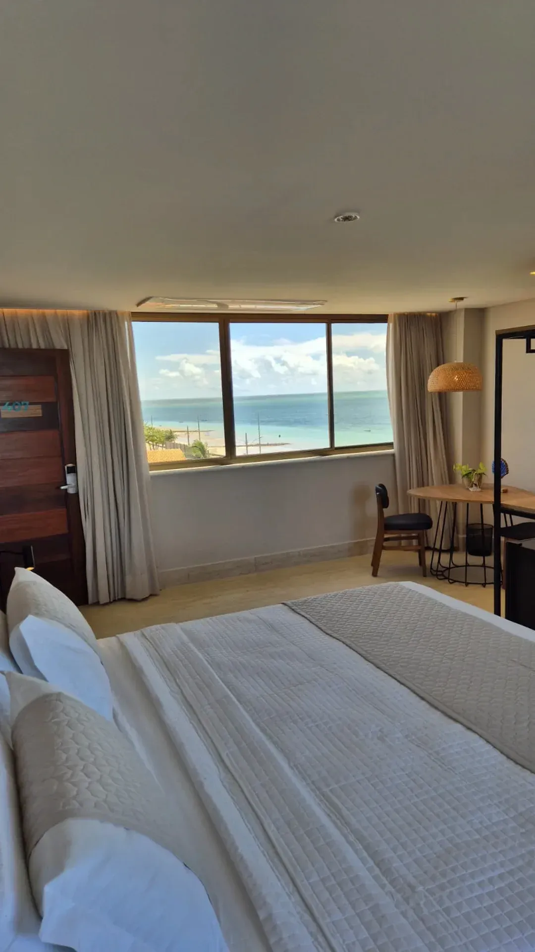 Quarto com colcha branca, janela aberta e vista para o mar.