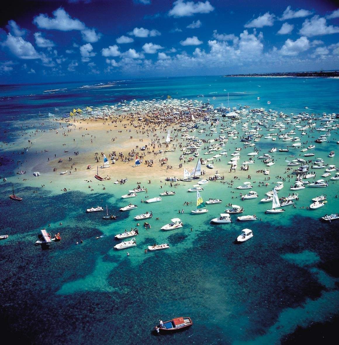 Vista aérea de um banco de areia lotado de barcos em um oceano azul-turquesa brilhante sob um céu azul.