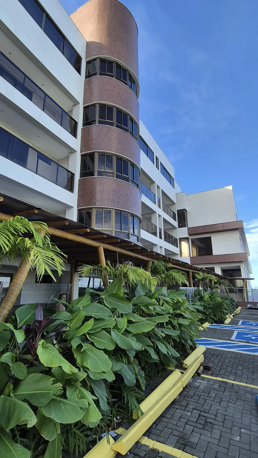 Fachada moderna do edifício com varandas curvas, vegetação exuberante e um caminho pavimentado sob um céu azul.