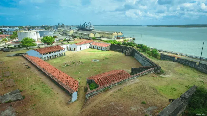 Vista aérea de um forte costeiro histórico com edifícios de telhados vermelhos junto ao mar sob um céu azul.