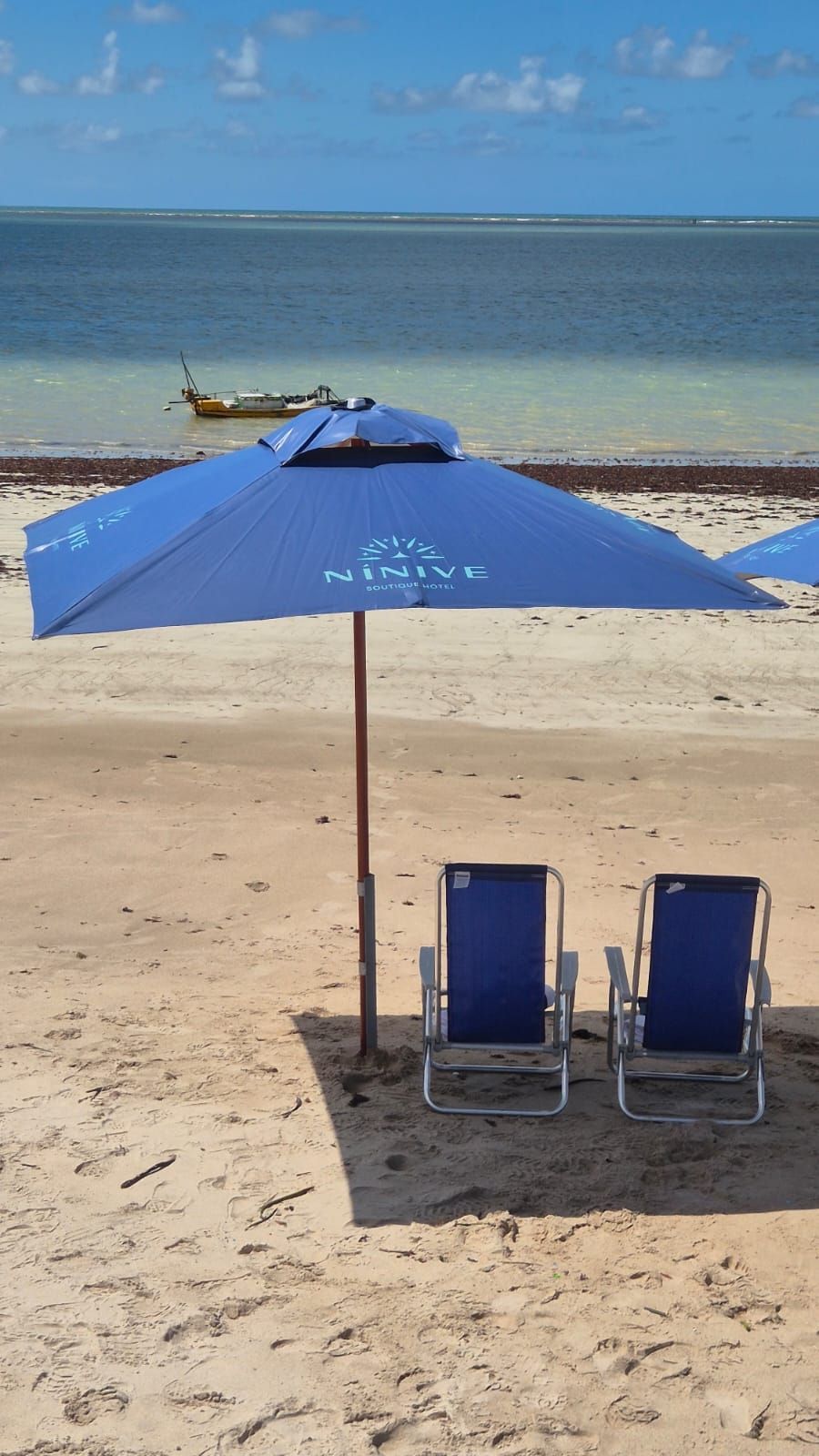 Guarda-sol azul com duas cadeiras azuis na areia, ao lado do oceano calmo.