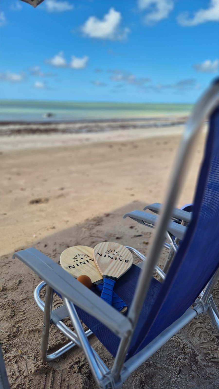 Cadeira de praia azul com remos em uma praia de areia com águas calmas e turquesas.