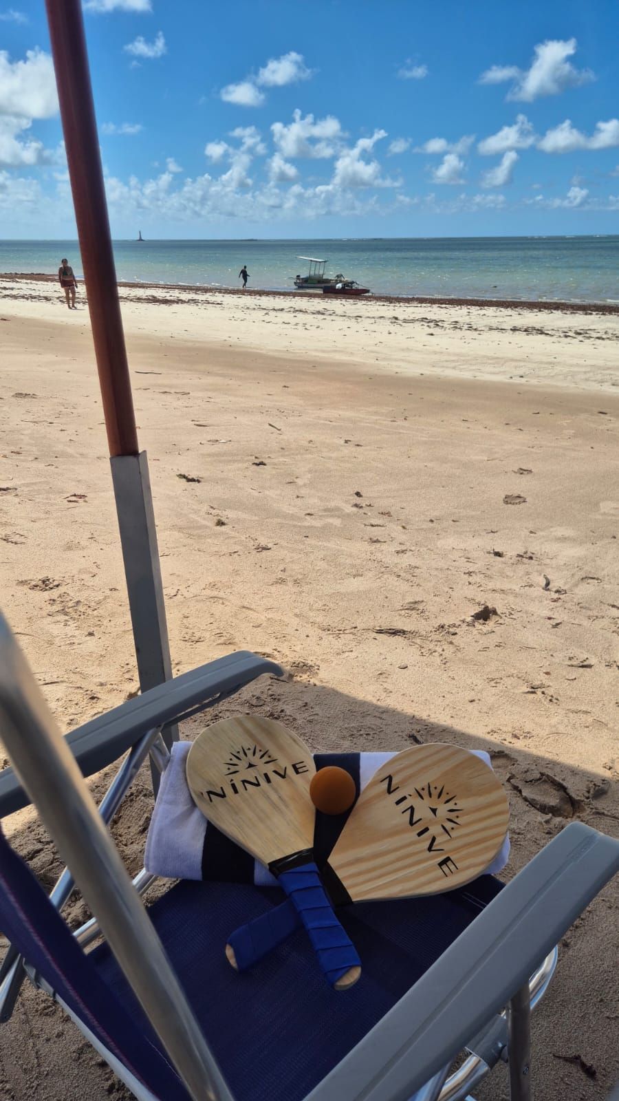 Cadeira de praia com dois livros abertos e um leque em uma faixa de areia, com o oceano e o céu azul ao fundo.