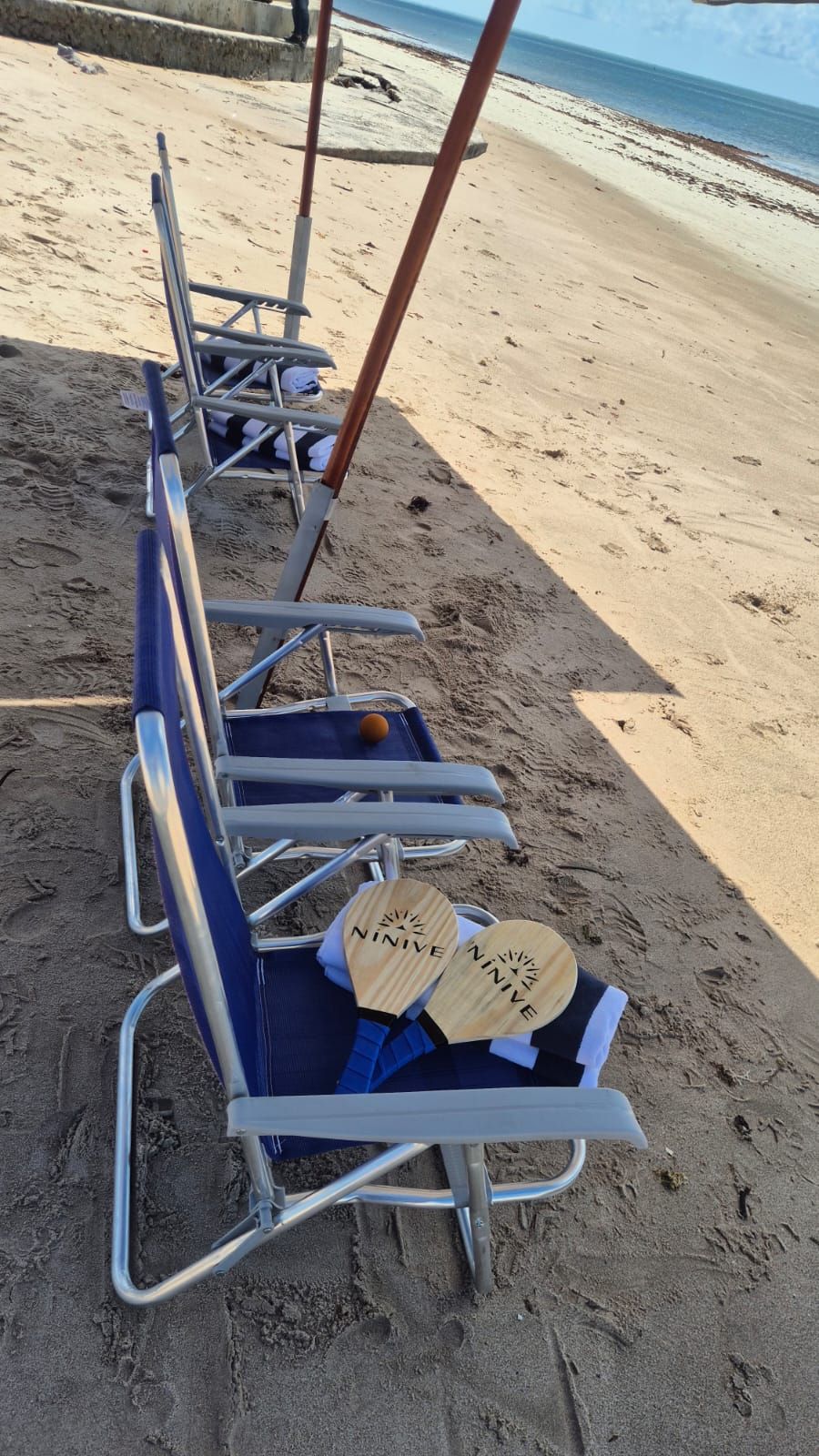 Cadeiras de praia azuis enfileiradas na areia, com um par de chinelos na cadeira mais próxima.
