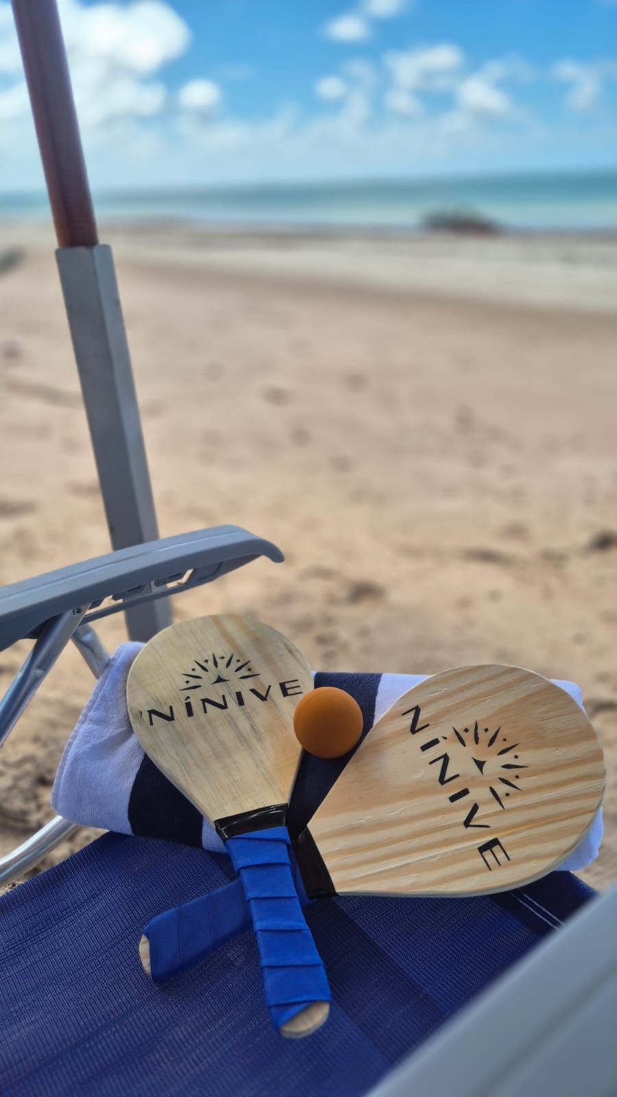 Cadeira de praia com dois remos de madeira e uma pequena bola sobre uma toalha azul, com o oceano ao fundo.