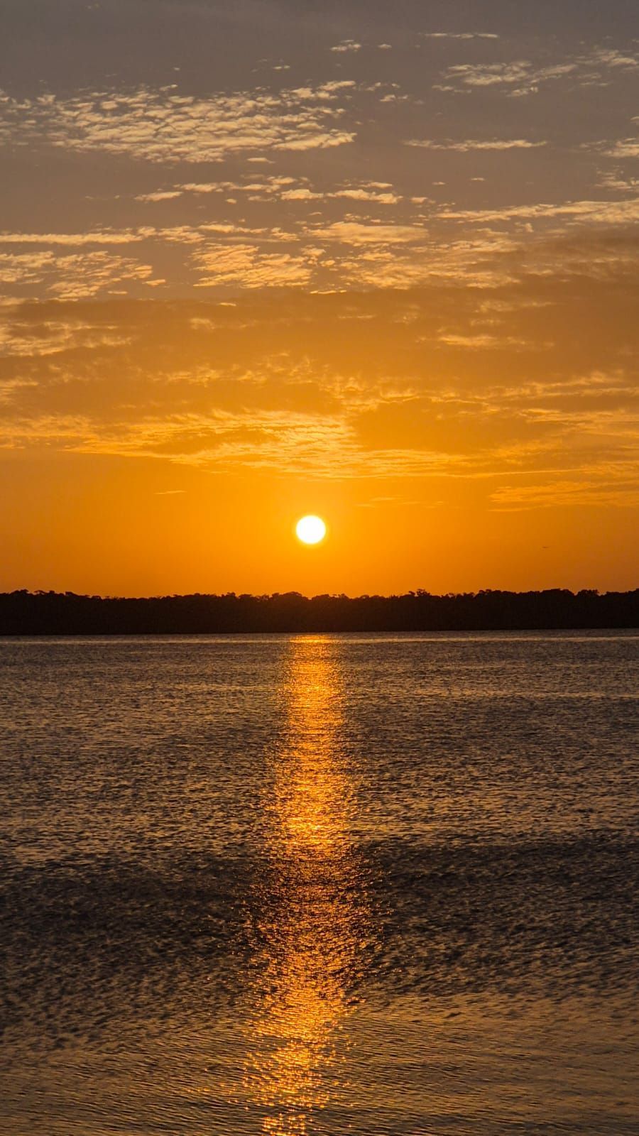 Pôr do sol dourado sobre um lago calmo, com a luz alaranjada refletida na água sob um céu com algumas nuvens.