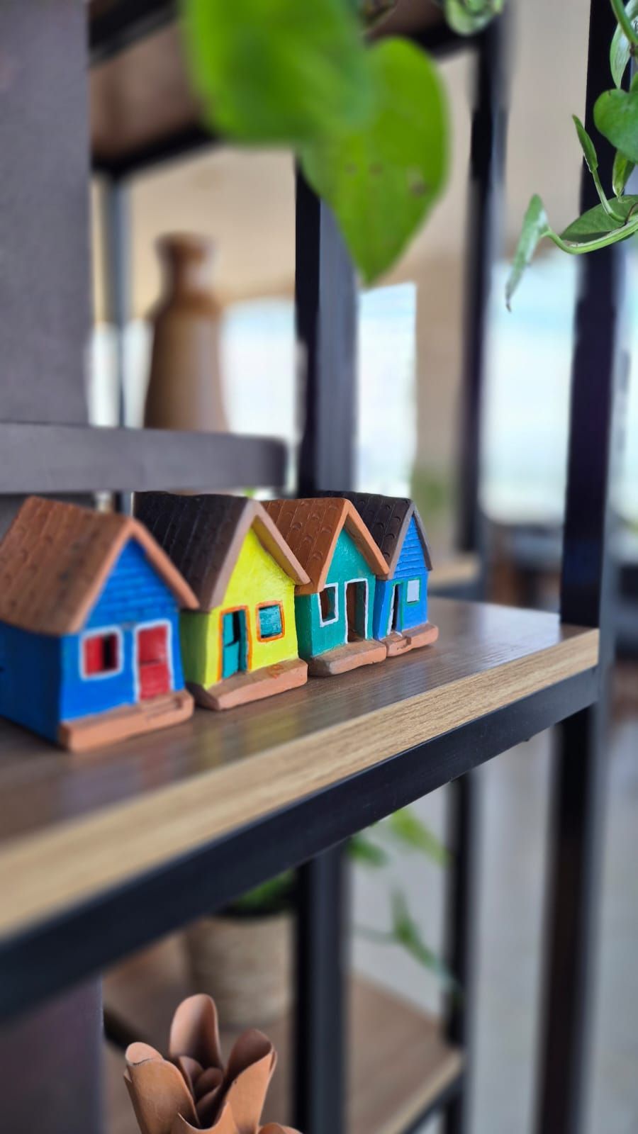 Casinhas coloridas em miniatura numa prateleira com plantas ao fundo.