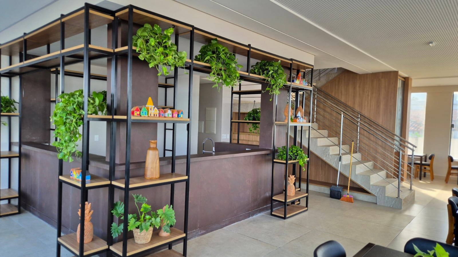 Hall de entrada moderno com estantes de metal preto, plantas suspensas, balcão de recepção e escadaria.