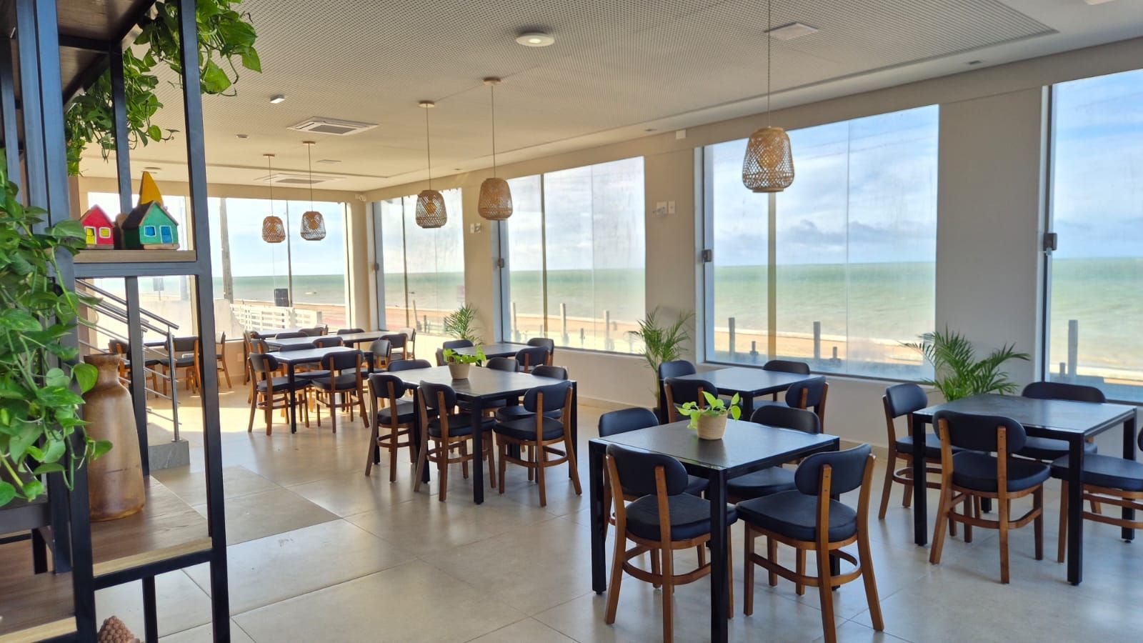 Café luminoso à beira-mar com mesas e cadeiras vazias e grandes janelas com vista para o mar.
