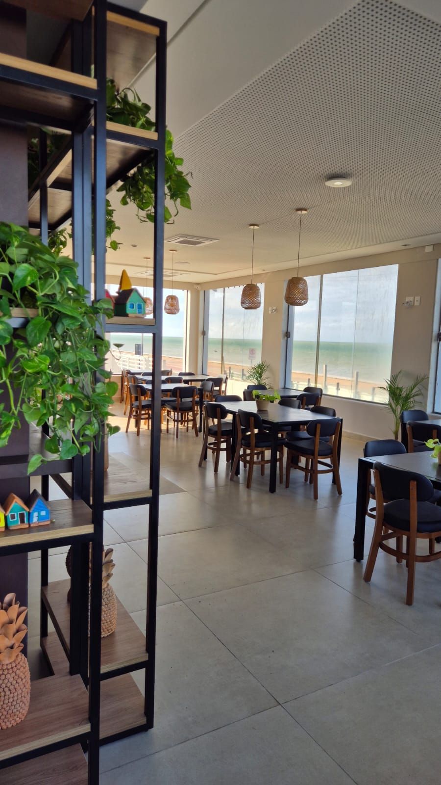 Interior luminoso de um café com mesas, cadeiras, plantas e grandes janelas com vista para o mar.