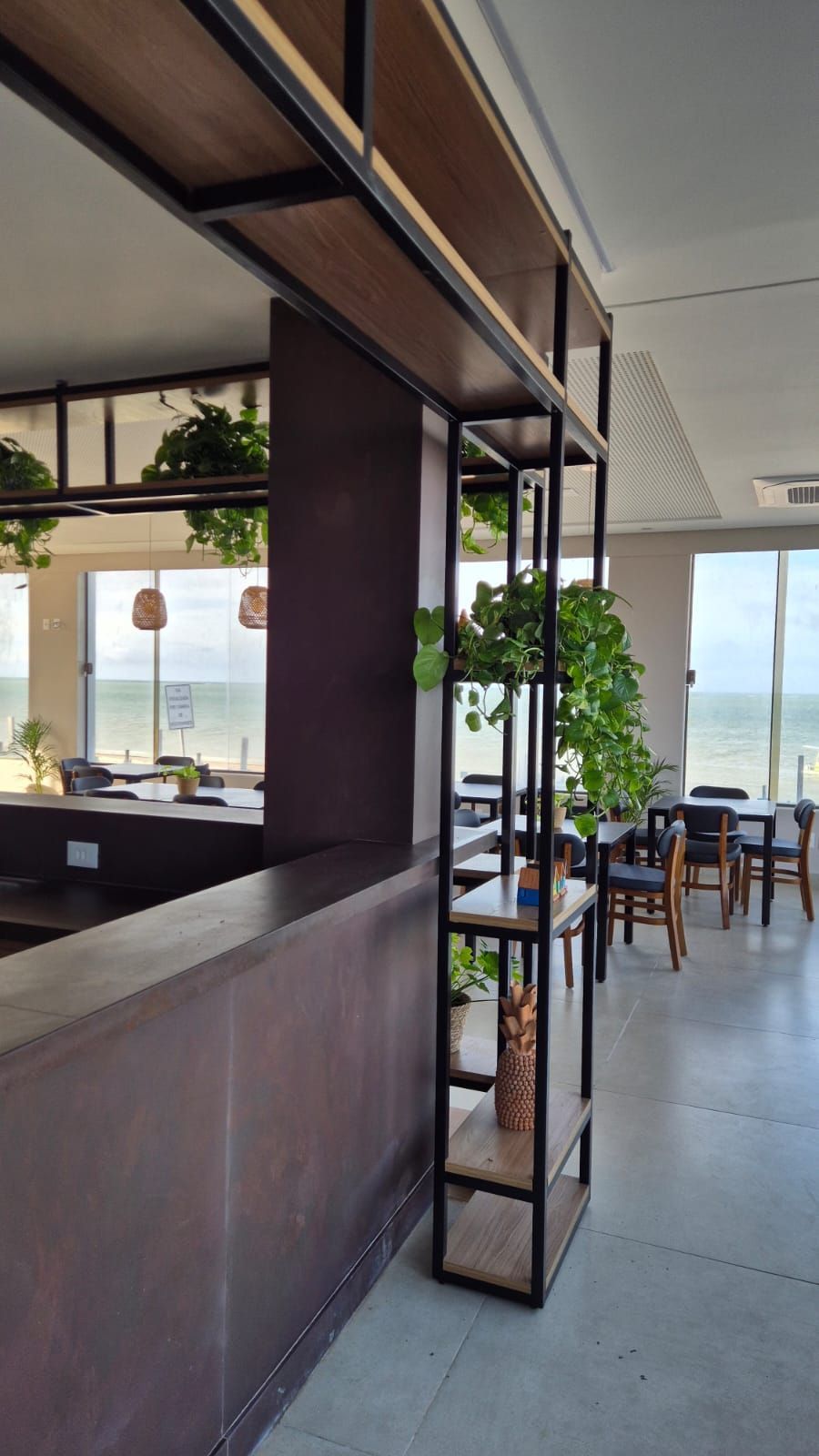 Interior de café moderno com balcão de madeira, mesas, cadeiras e grandes janelas com vista para o mar.